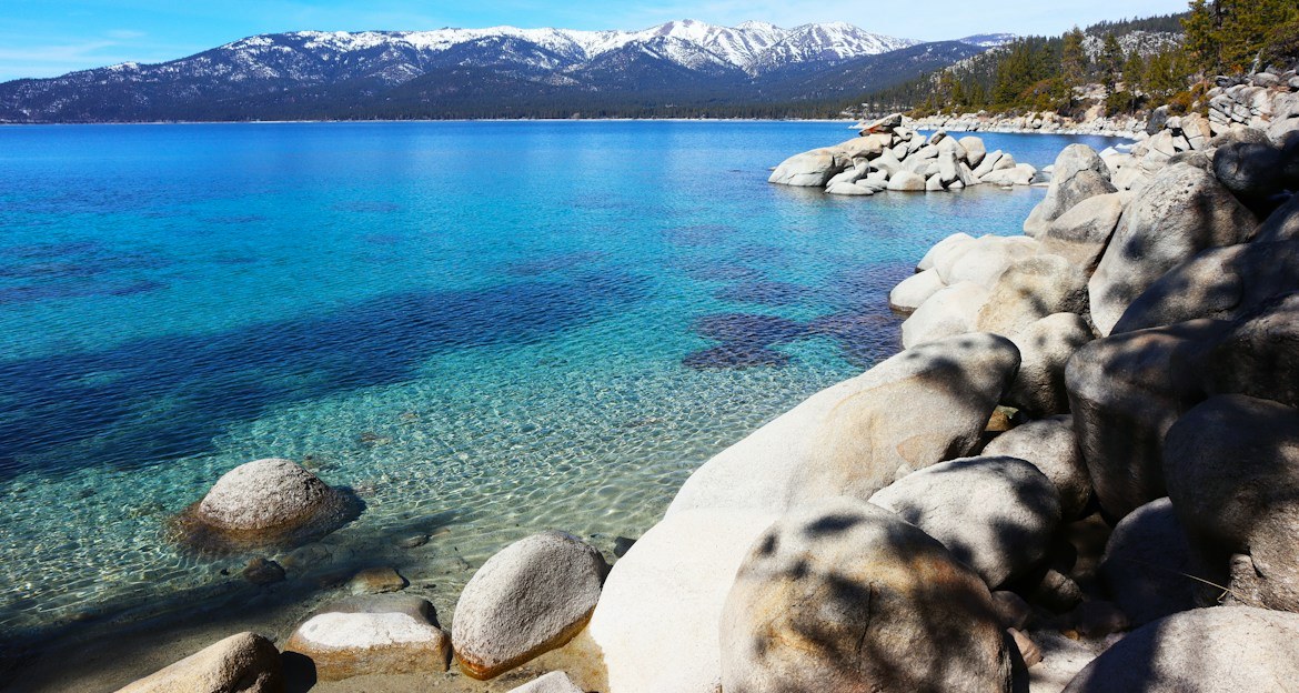 Clear blue water and granite boulders along the Lake Tahoe shoreline with snowy peaks